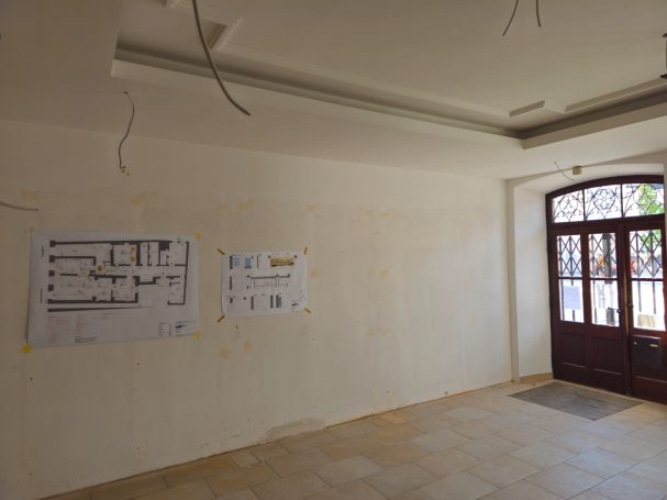 Leerer Raum mit Bauplänen an der Wand und einem Fenster mit Holztür.